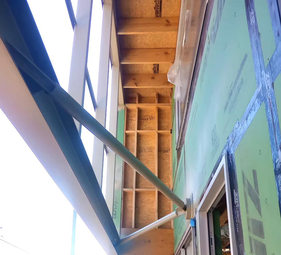 Steel awning strut and outrigger bearing on green ZIP-sheathed wood framing, photographed from below on site